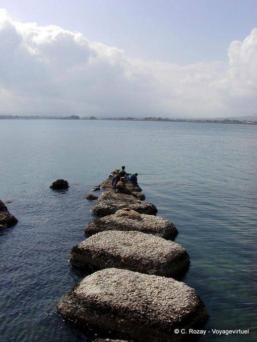 Fischer auf dem Pier in schwarzen Vulkansteinen, Syrakus, Sizilien - Italien
